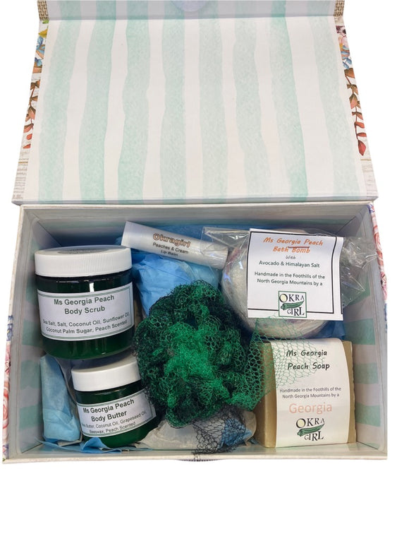 Top folding floral themed box lined with tissue paper containing small plastic containers of body scrub, body butter, a hand cut bar of peach soap wrapped with a thick strip of paper printed with Okra Girl's branding, standard tube of peach flavored lip balm, a small bath bomb wrapped in plastic, and a mesh exfoliating scrub